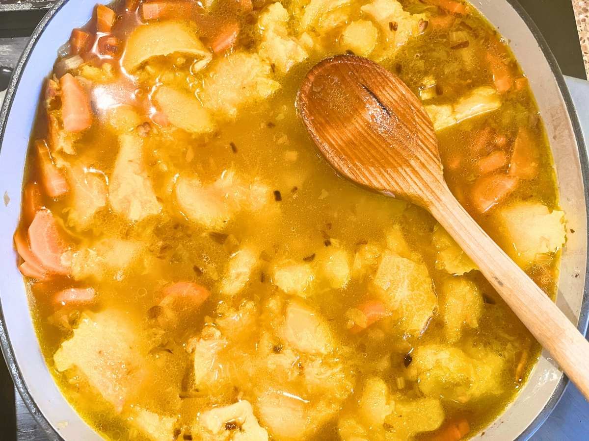 There are vegetables and butternut squash inside of a large pot with broth. There is a wooden spoon resting on top.