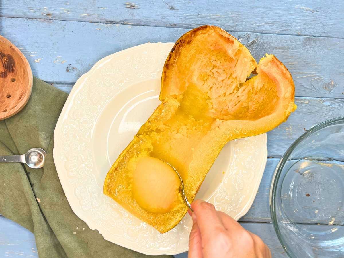 A woman is scooping out the flesh from a cooked butternut squash.