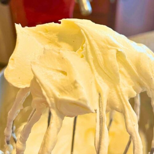 A woman is holding up a large whisk with cream cheese icing in it.