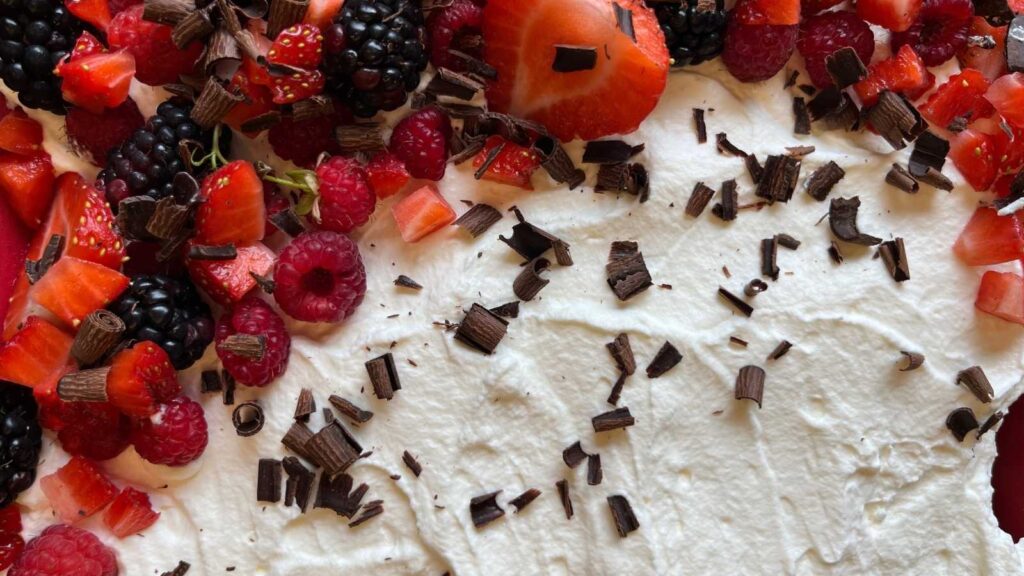 A tres leches cake in a red casserole dish. It is covered in berries and chocolate curls.