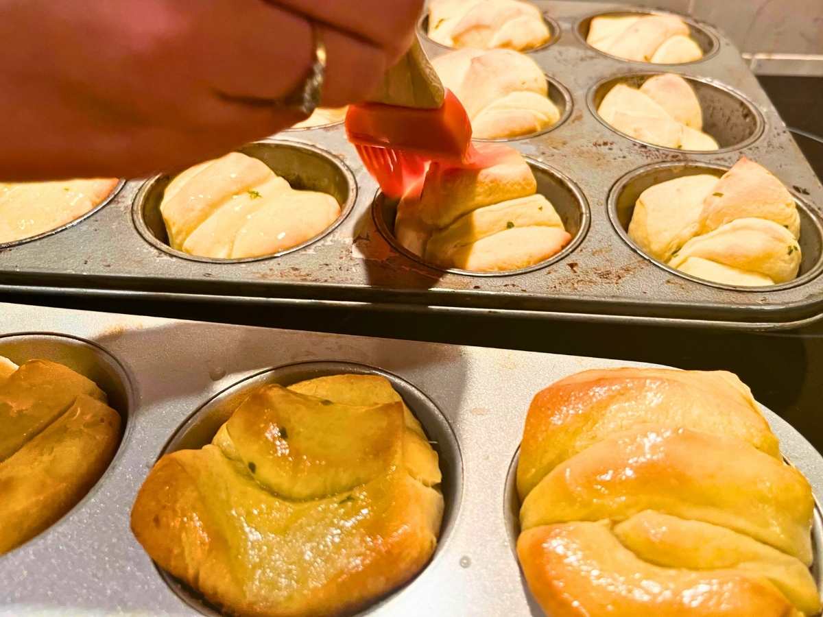 There is a woman brushing butter onto baked dinner rolls.