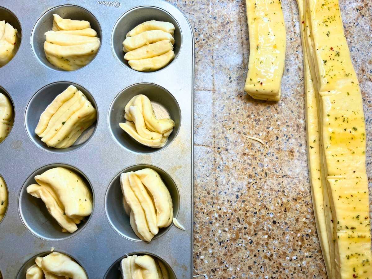 A muffin tin full of dough. There are garlic butter brushed strips of dough beside.