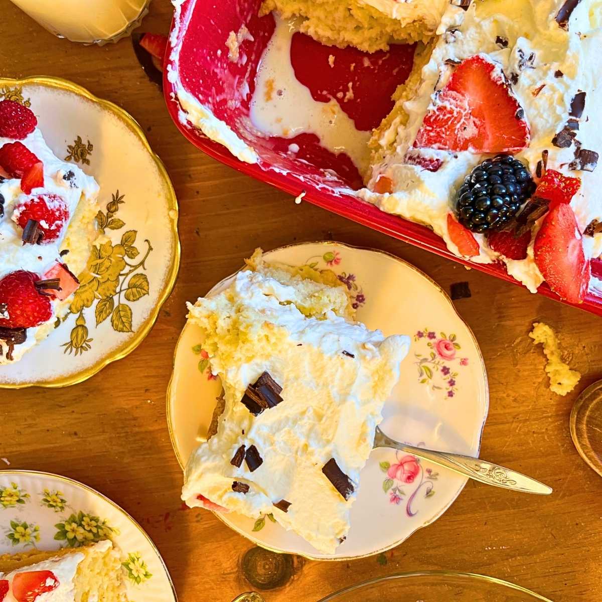 A cut tres leches cake in a red casserole dish. There are slices of cake on plates in front.