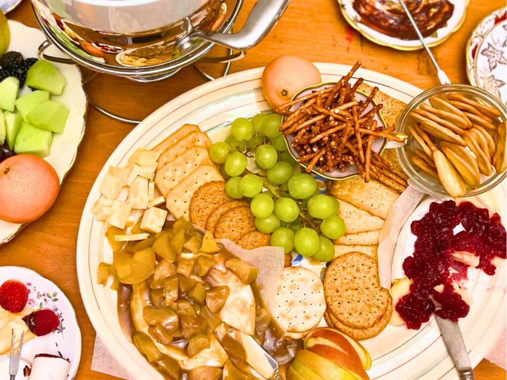 A large cheese platter with baked Brie, accompaniments, fresh, fruit, and plates of to the side.