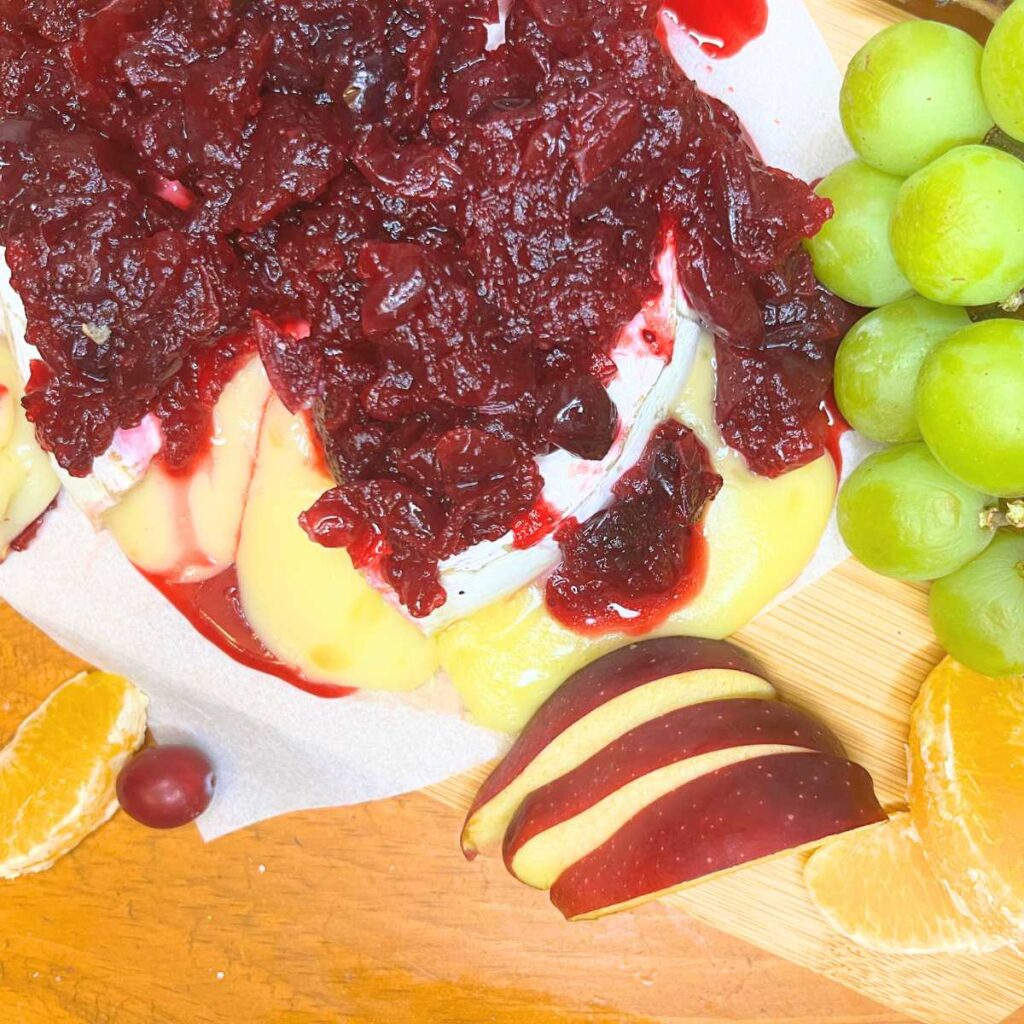 A baked brie with cranberry sauce on top. There is fresh fruit off to the side.