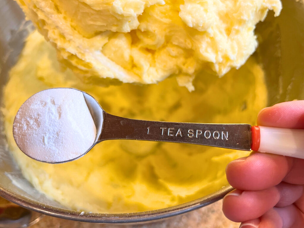 Creamed butter and sugar in the bowl of a stand mixer. A woman holding baking powder above the bowl in a measuring spoon.