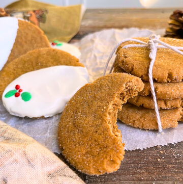 Gingerbread cookies sitting on top of a pieces of parchment paper on a wooden table. Some cookies are decorated with white chocolate and Christmas sprinkles, others are stacked and tied with kitchen twine. One has a bite out of it.