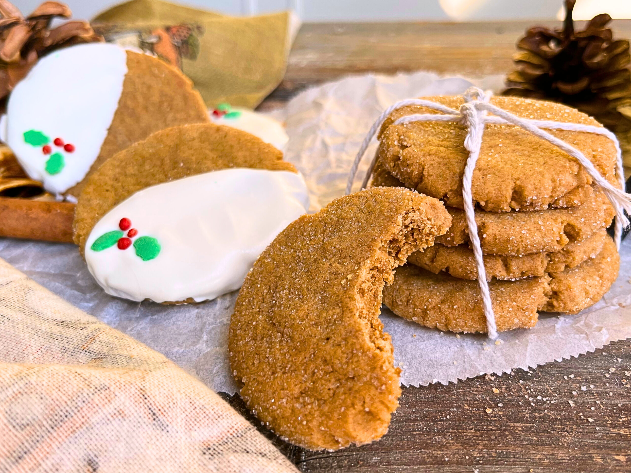 Gingerbread cookies sitting on top of a pieces of parchment paper on a wooden table. Some cookies are decorated with white chocolate and Christmas sprinkles, others are stacked and tied with kitchen twine. One has a bite out of it.