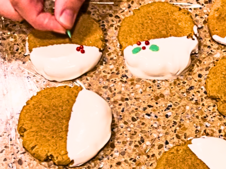 Woman placing red and green holly sprinkles on to a gingerbread cookie with white chocolate.