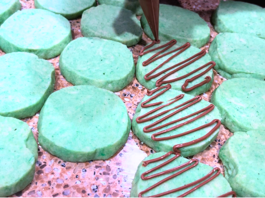 Woman using a piping bag to drizzle melted chocolate onto mint Christmas cookies.