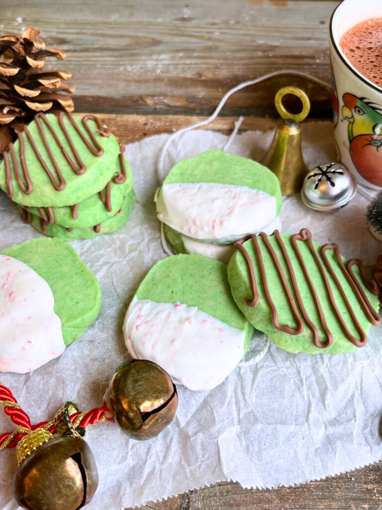 Mint Christmas cookies on parchment paper on a wooden table. There are bells around them and a mug of hot chocolate in the background.