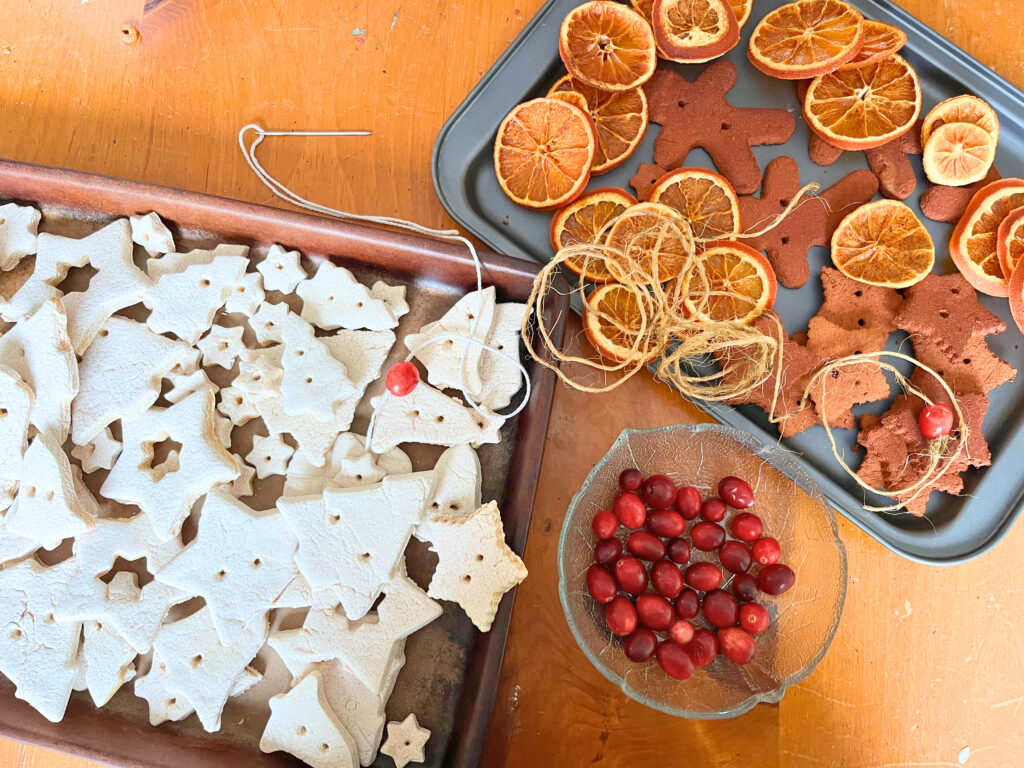 A tray of salt dough Christmas shapes. Another tray with cinnamon dough shapes and dried orange slices. A small glass bowl with cranberries. A string of kitchen twine with a needle and a cranberry on the end, and a length of jute twine with a cranberry on the end.
