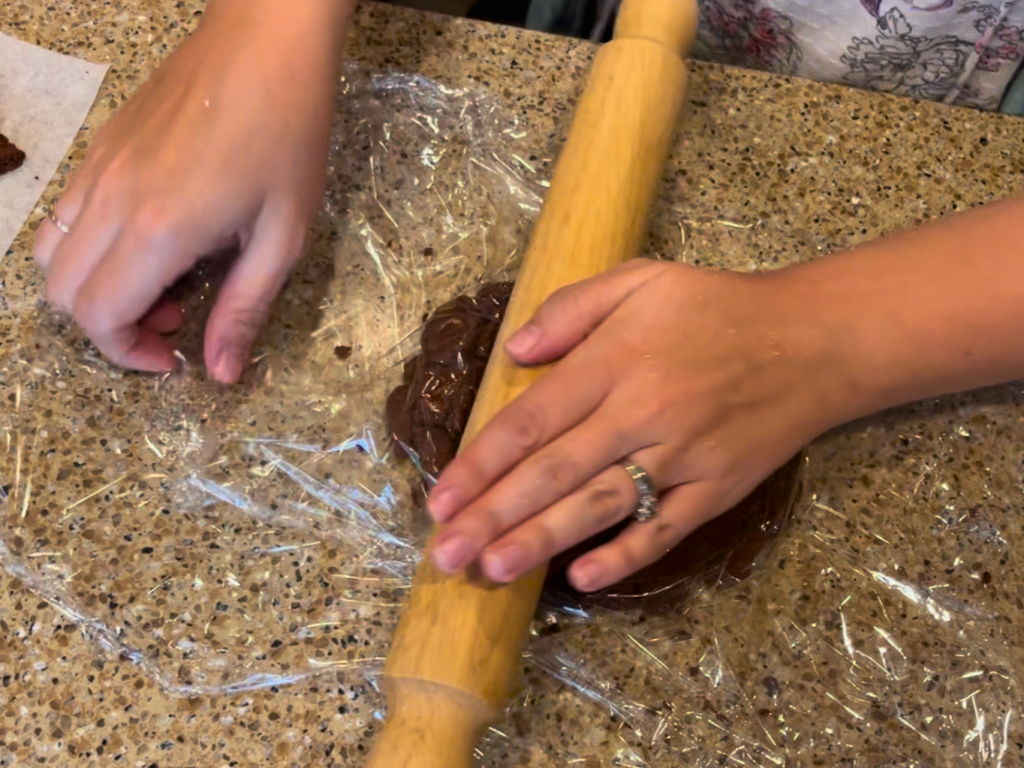 Woman rolling out cinnamon dough in between two pieces of cling-film, with a wooden rolling pin.