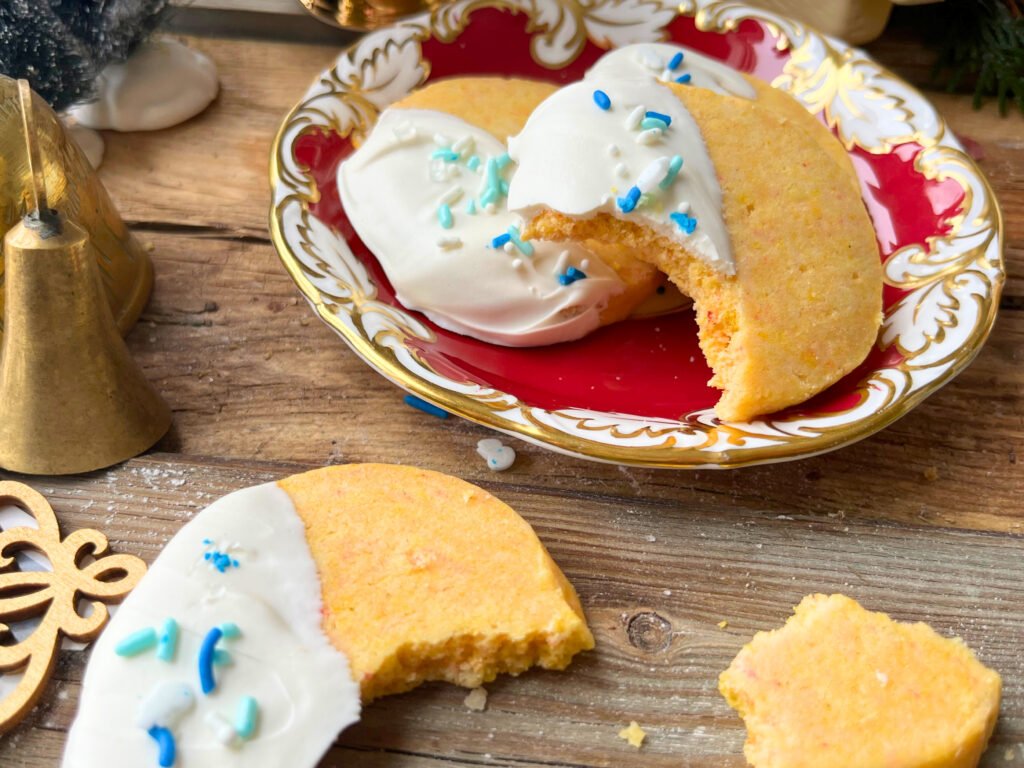 A red plate with orange cardamom shortbread cookies. They have white chocolate and blue sprinkle decorations.