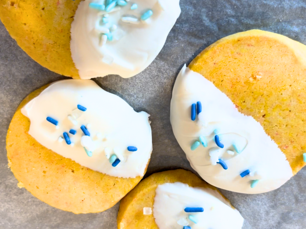 Four orange cardamom shortbread cookies dipped in white chocolate and decorated with blue sprinkles.