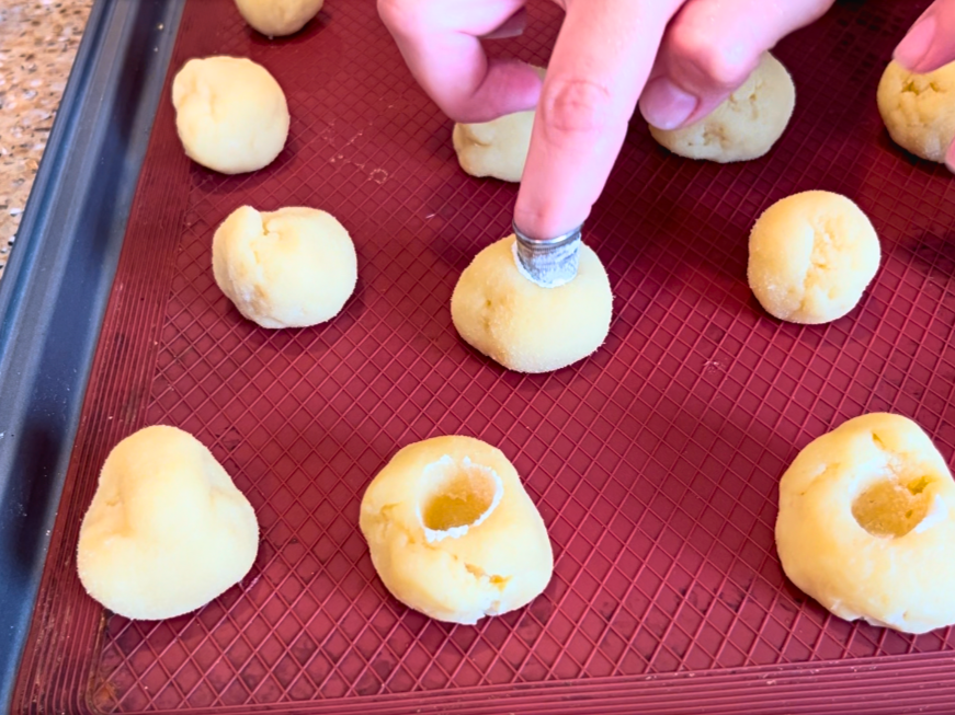 Woman using a thimble to press depressions into the centre of dough balls on a baking sheet.