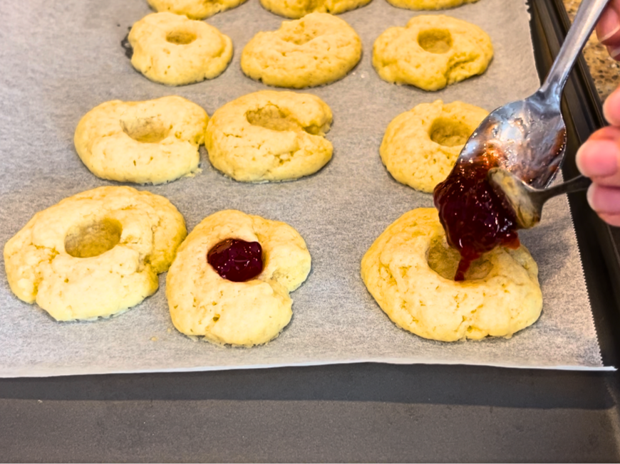 Woman using spoons to fill depressions in thumbprint cookies with jam.