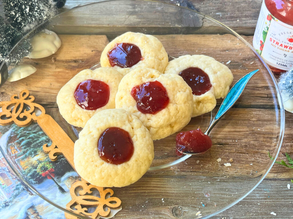 A glass plate with five thumbprint cookies on it. A jam jar in the background and a small blue spoon with jam on it on the plate.