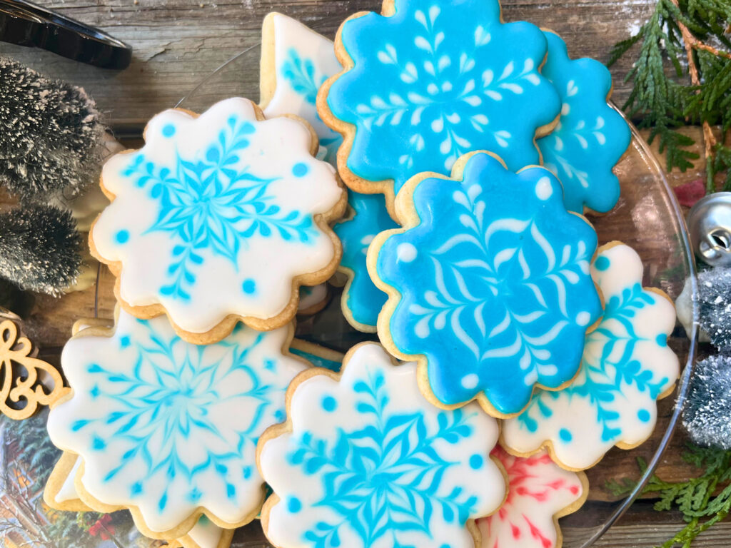 A glass plate with decorated snowflake sugar cookies with royal icing in white and blue, one in red and white.