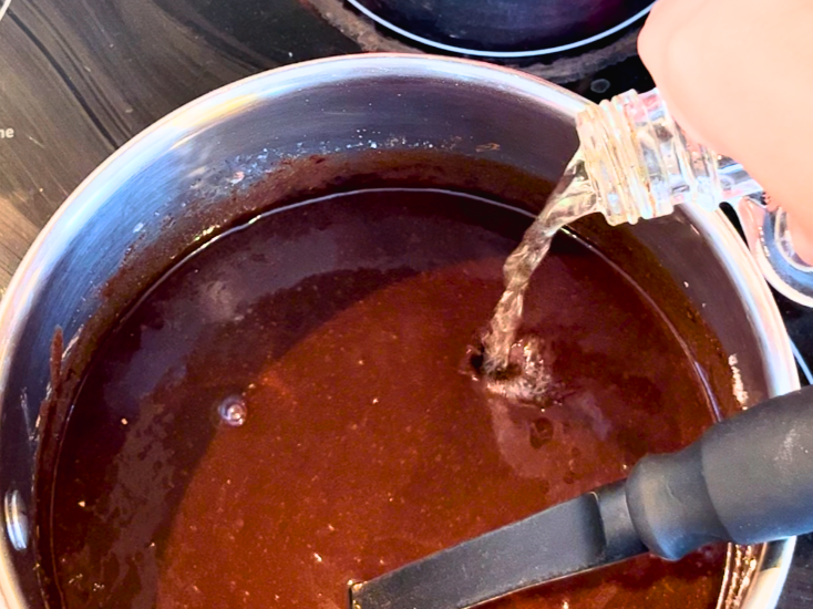 Woman pouring vanilla extract into a sauce pot with a chocolate mixture. 