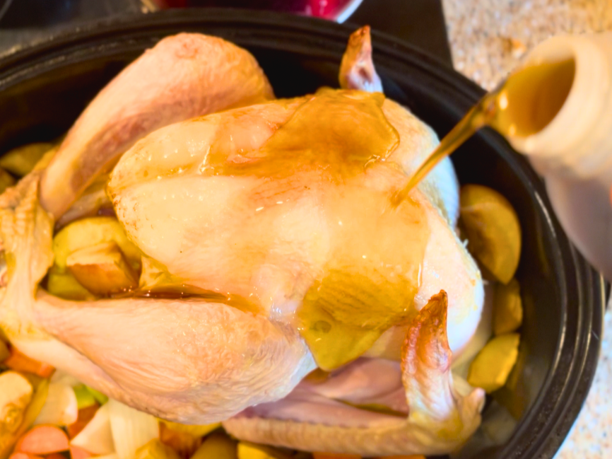 An uncooked turkey sitting on a bed of fruit and vegetables. Maple syrup is being poured over top of it.