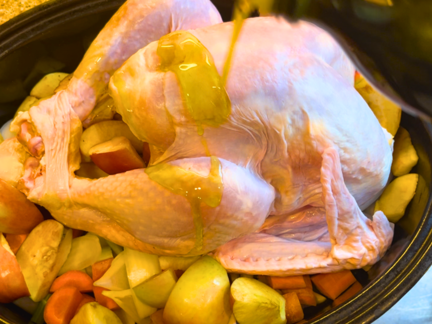 An uncooked turkey sitting on a bed of cut fruit and vegetables. Oil is being drizzled over top of it.