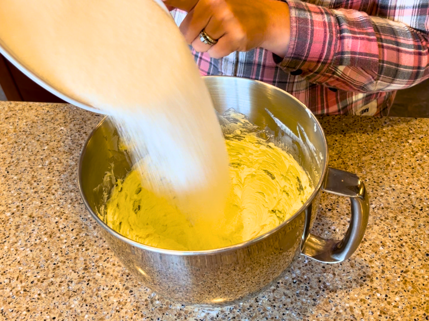Woman pouring sugar into a bowl of creamed butter.