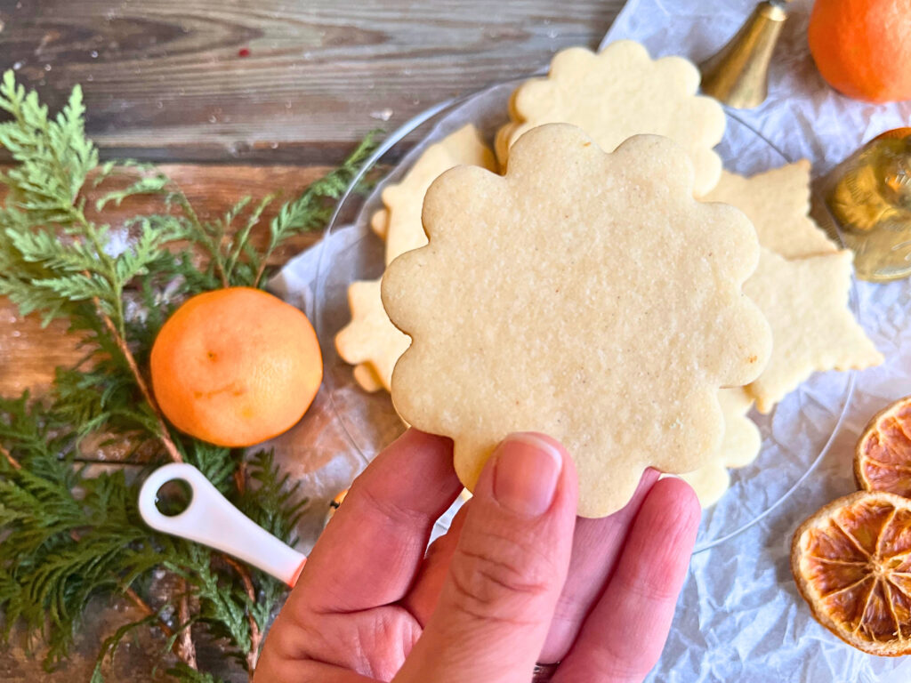 A woman holding an orange and cardamom sugar cookie. A glass plate with more sugar cookies in the background.