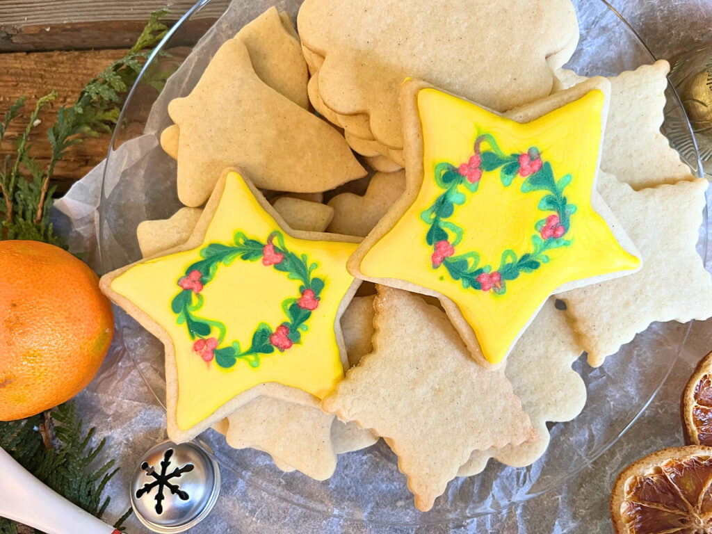 A glass plate with orange and cardamom sugar cookies on it. Two of them are decorated starts with royal icing wreaths on them.