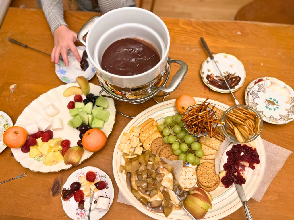 A cheese platter with crackers, pretzels, apples, and grapes. Two wheels of baked brie, one with apples and caramel and one with cranberry sauce. A chocolate fondue in the background and a fruit tray.