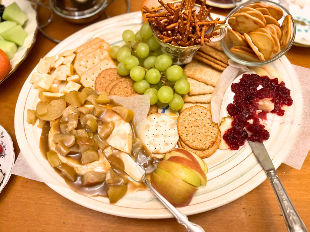 A cheese platter with baked brie with apple caramel and one with cranberry sauce. Crackers, pretzels, sliced apples and grapes also on the tray.
