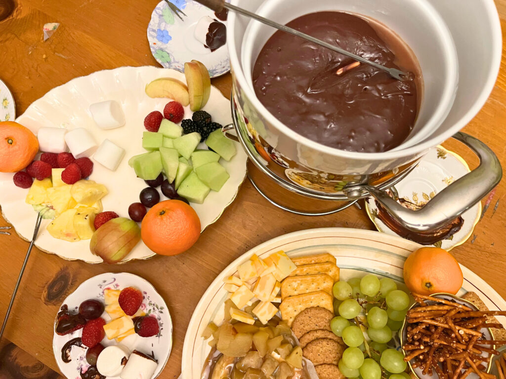 A chocolate fondue set up with a fruit platter off to the side. There is a cheese platter with fruit cheese and crackers in the foreground.