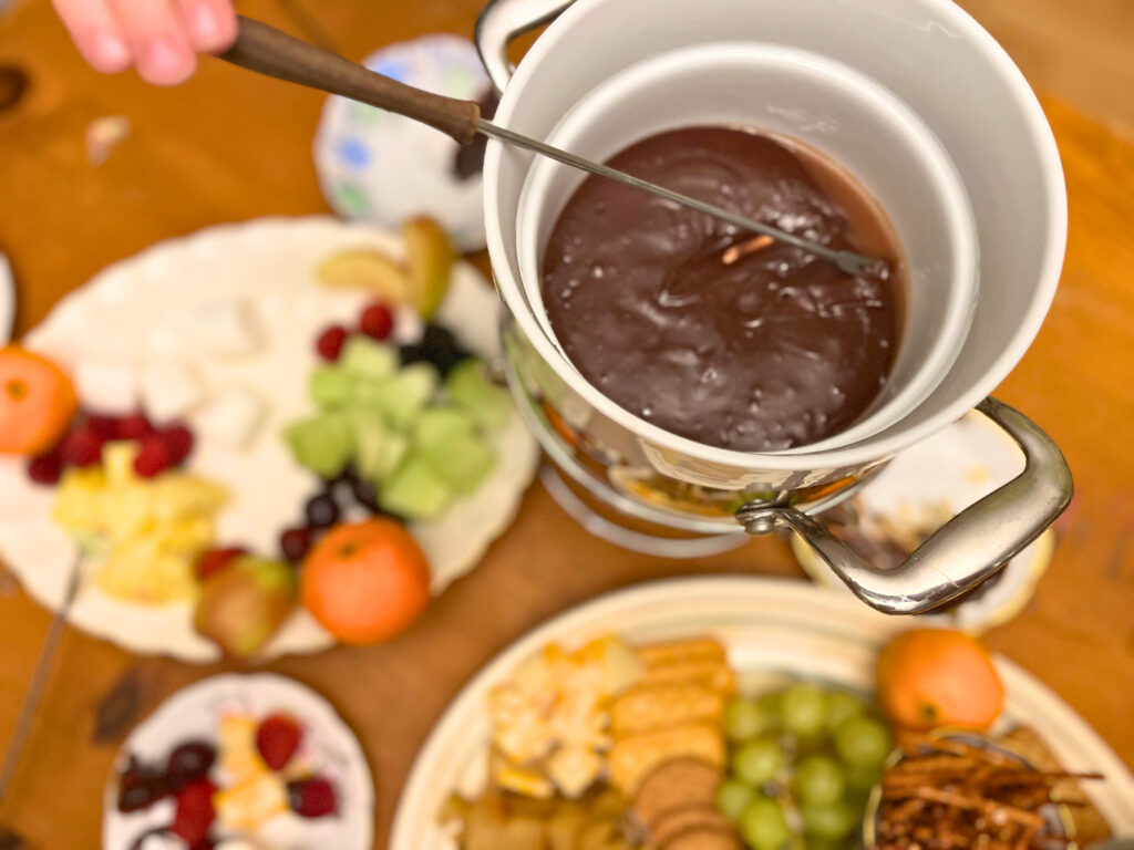 Chocolate fondue with a plate of dippers on the side and a cheese platter in the foreground.