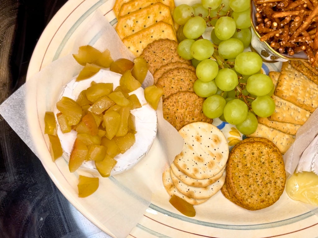Baked brie with cooked apples on top. Crackers and grapes are on the platter beside it.