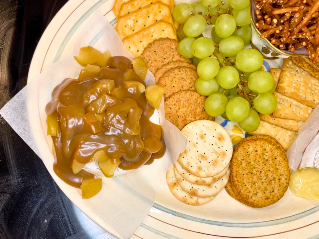 Baked brie with apples and caramel on top. Crackers and grapes are on the platter beside it.