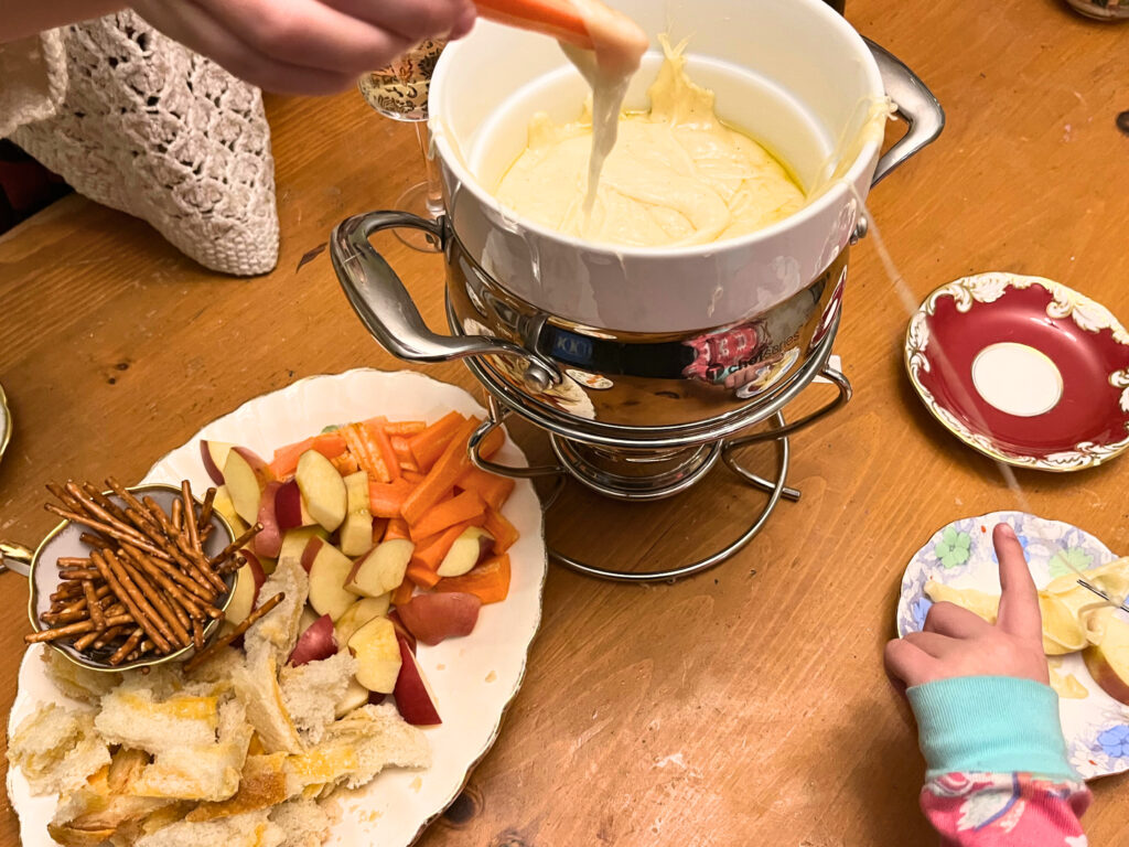 A pot of cheese fondue with a plate of dippers on the side. Two girls are using the fondue pot.