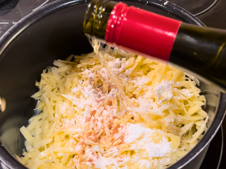 Grated cheese in a sauce pot with cornstarch, nutmeg, and salt and pepper. White wine is being poured inside.