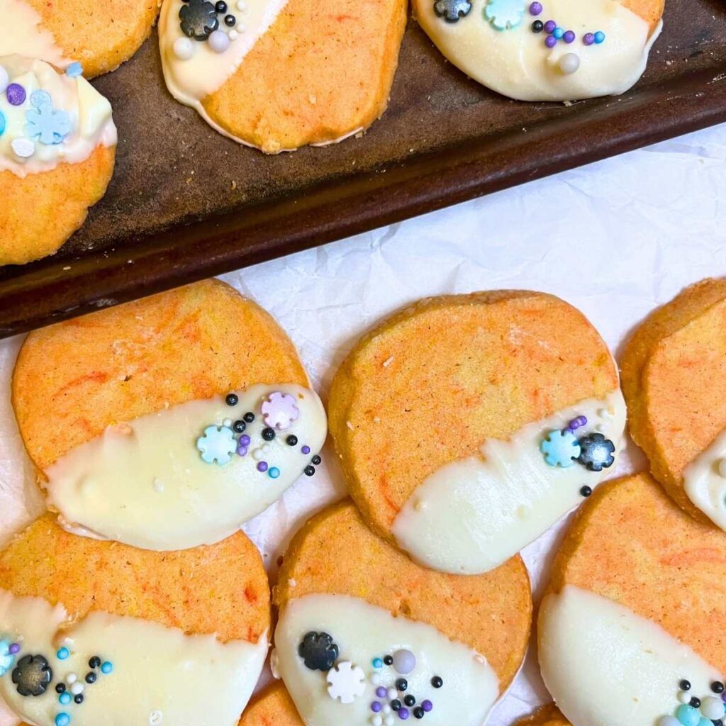 Some orange and cardamom cookies that are decorated in white chocolate and sprinkles. Some are on a cookie sheet and some are on white parchment paper.