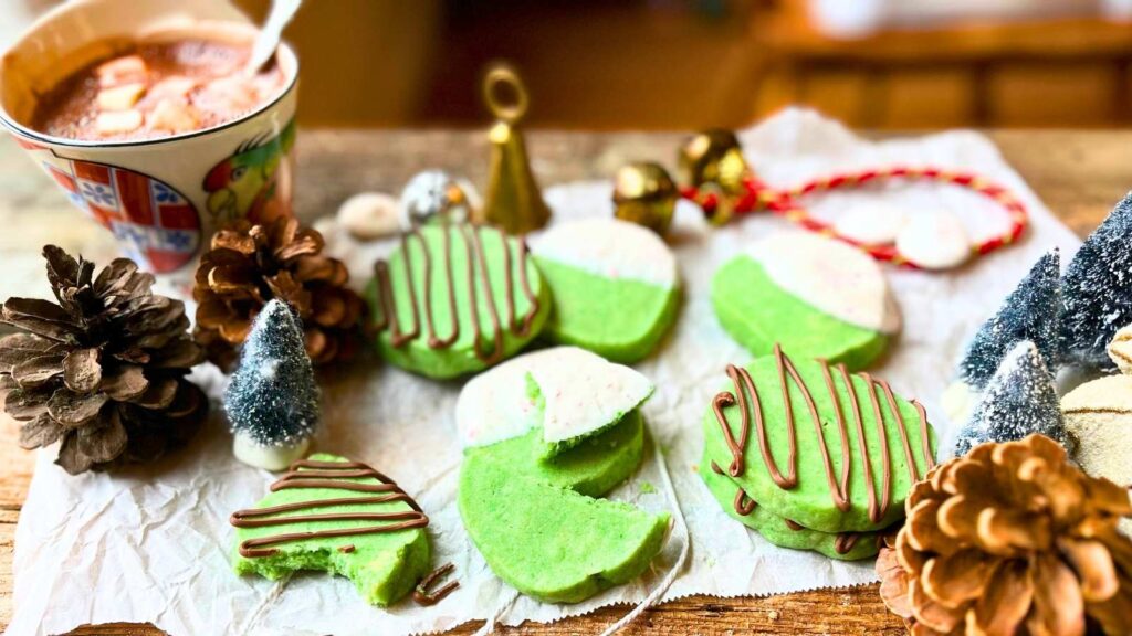 Some mint shortbread cookies that are decorated with chocolate on a piece of parchment paper. There is a mug of hot chocolate off to the side.