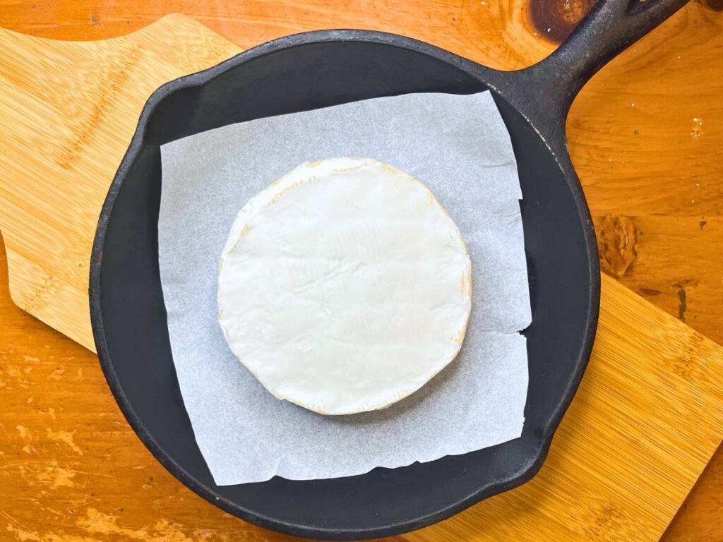 A wheel of Brie cheese on a piece of parchment paper in a cast iron skillet.