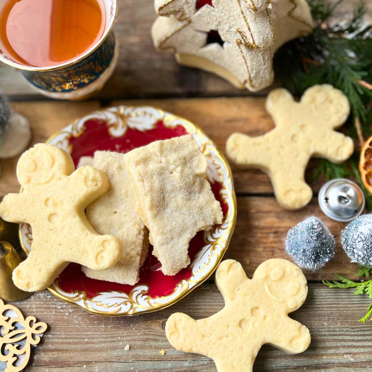 Some shortbread cookies on a red plate. Some are in the shape of gingerbread men. There is a cup of tea in the background.