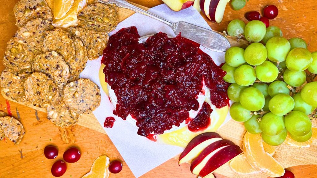 A wooden platter with a baked Brie covered in cranberry sauce. Thre are crackers and fresh fruit to the side.