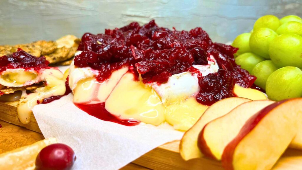 A baked Brie with cranberry sauce on top. It is on a wooden cutting board with sliced apples.