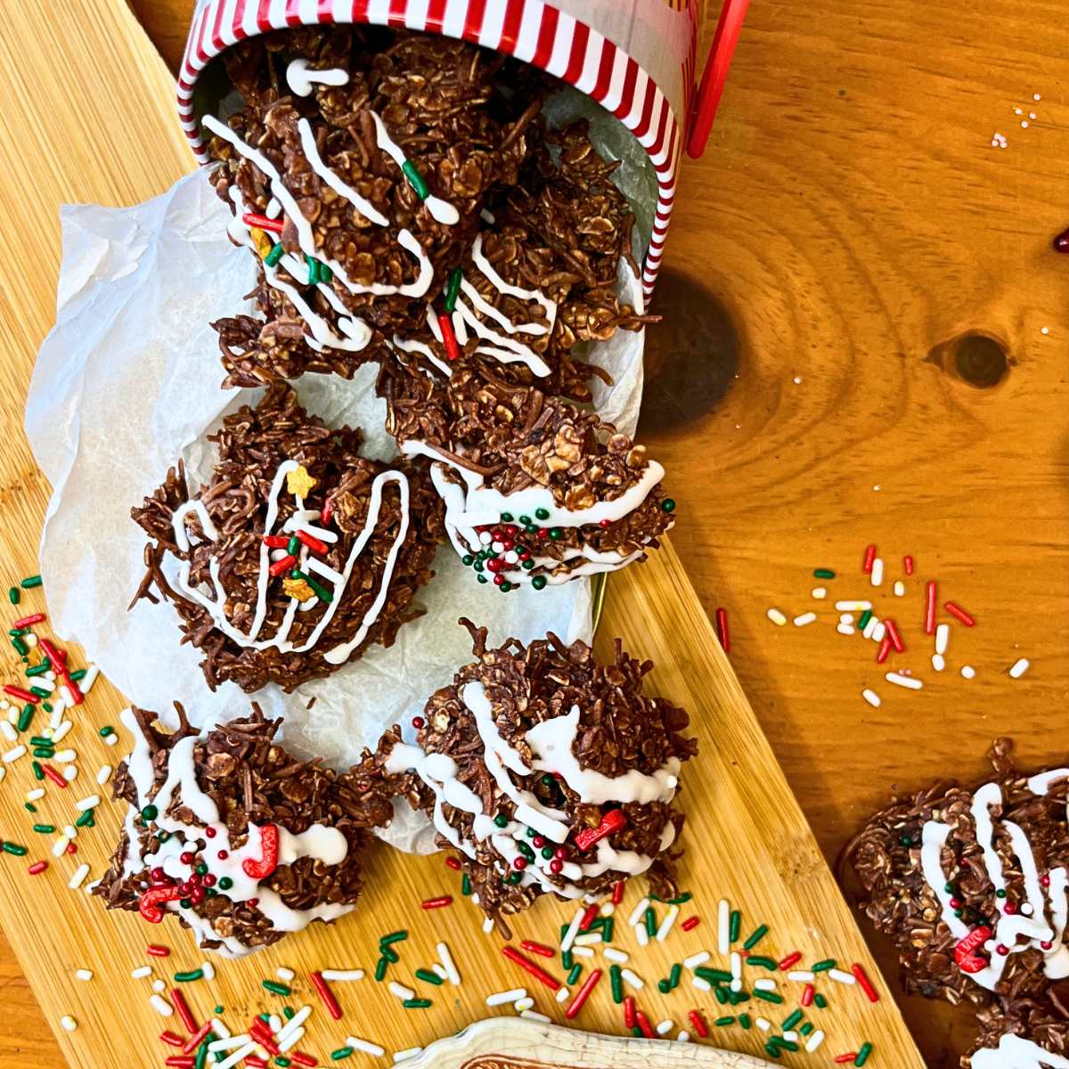 Some chocolate haystack cookies decorated with white icing and sprinkles. they are in a Christmas cookie tin.