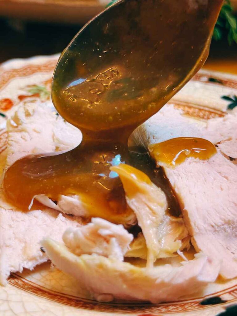 Sliced turkey on a plate. A woman is spooning gravy onto it.