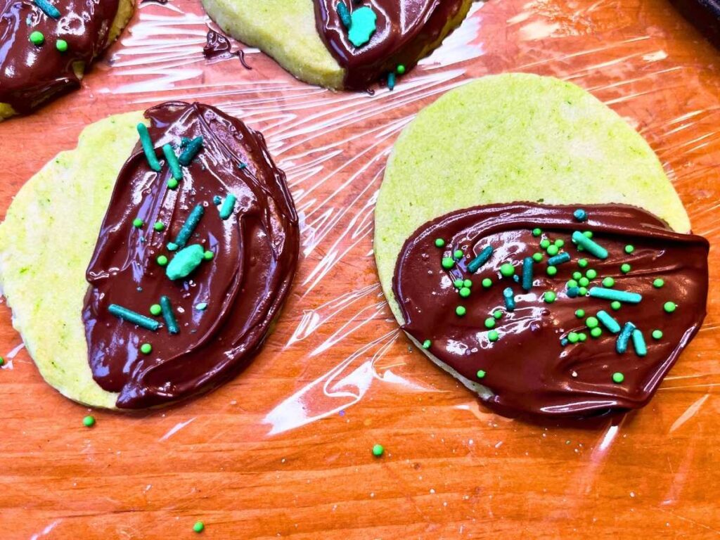 Some mint shortbread cookies on a piece of plastic wrap. They have been dipped in chocolate and have sprinkles on top.