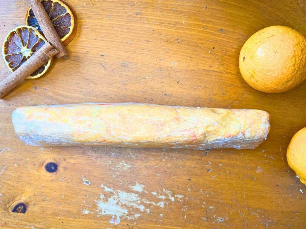 A log of orange and cardamom shortbread dough wrapped in plastic wrap.