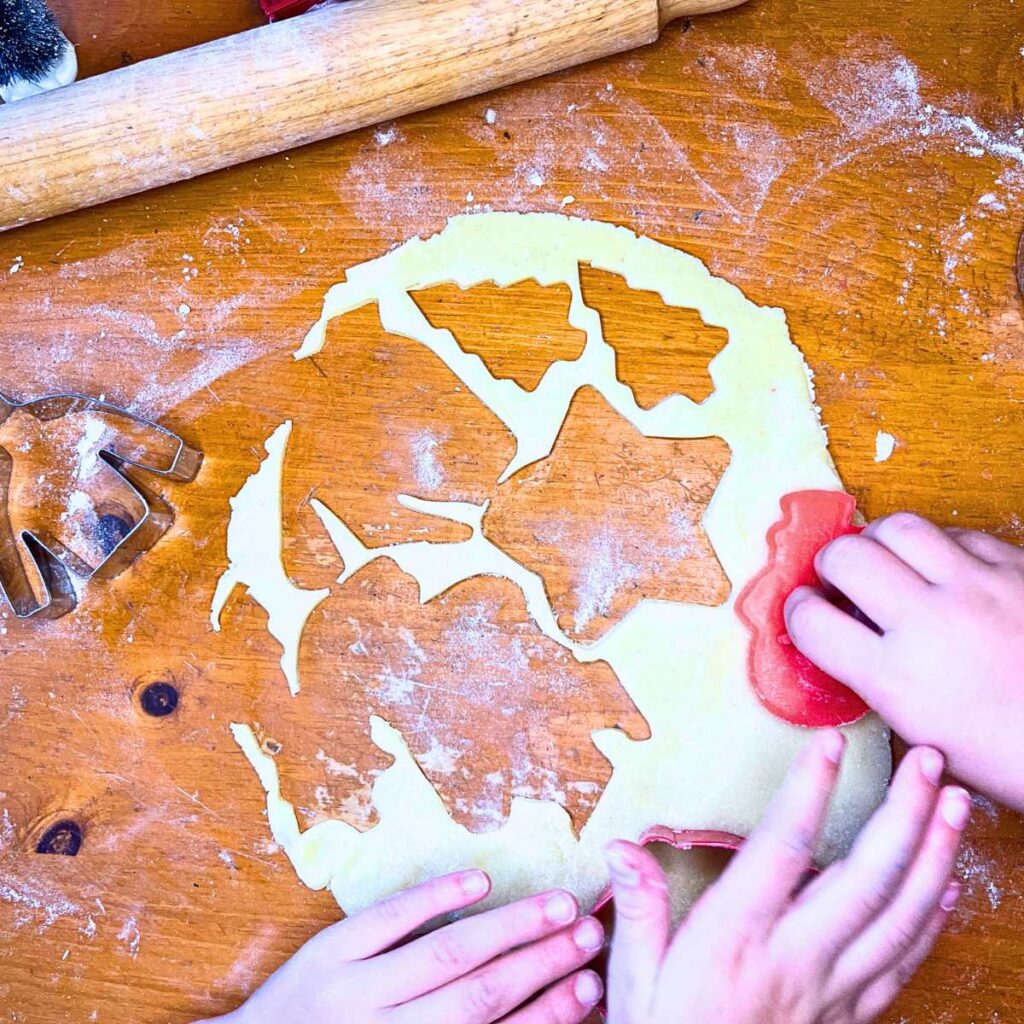 Some children's hands cutting sugar cookie dough with cookie cutters.