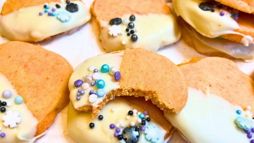 Orange cookies that have been dipped in white chocolate and decorated with sprinkles. One has a bite out of it.