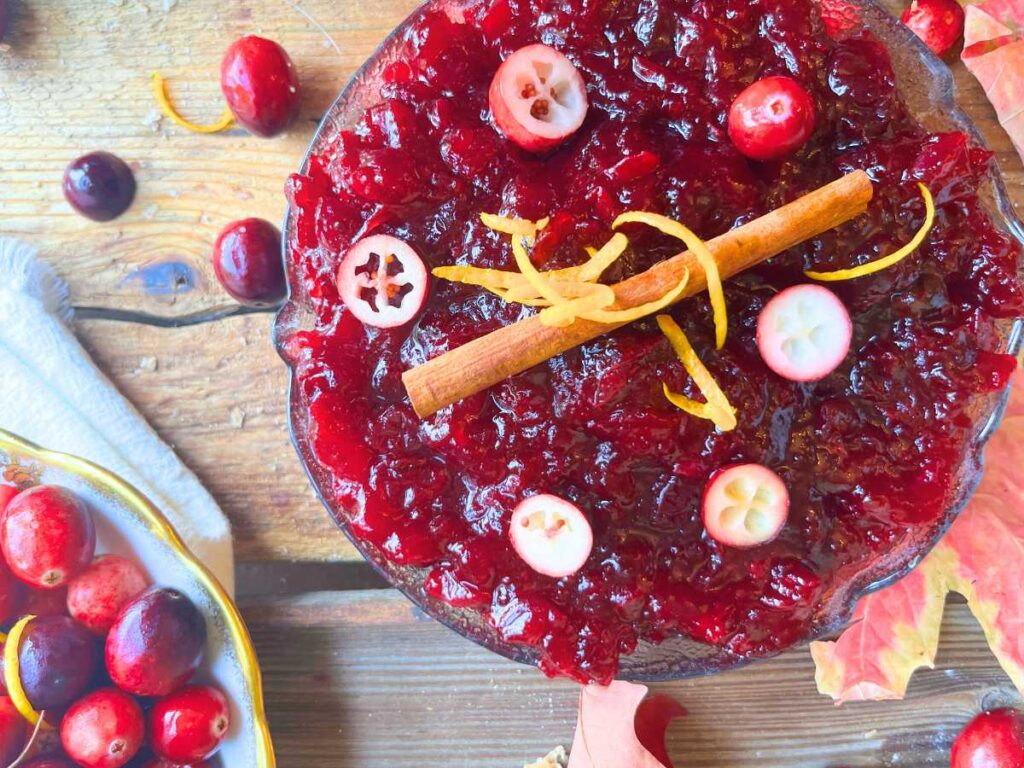 A bowl of cranberry sauce in a glass bowl. There are sliced cranberries on top, a cinnamon stick, and orange zest.